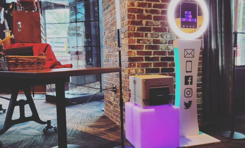 Selfie photo booth with an illuminated ring, touchscreen, and printer beside a brick wall