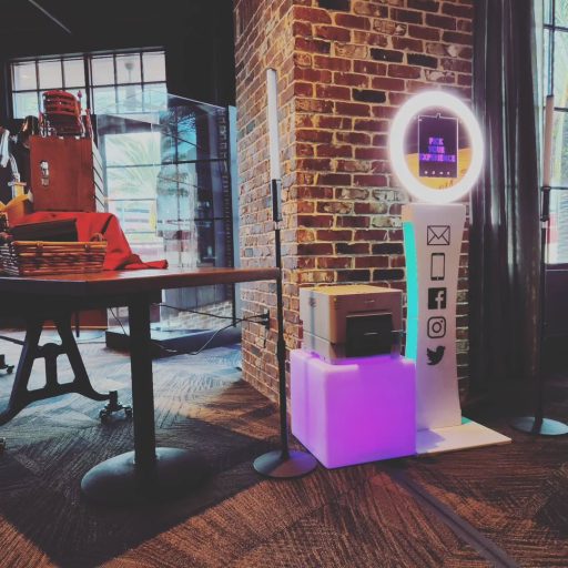 Selfie photo booth with an illuminated ring, touchscreen, and printer beside a brick wall